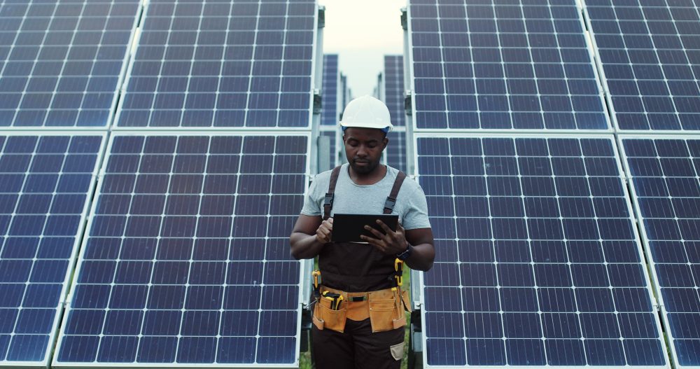 Portrait of african american electrician engineer in safety helmet and uniform using tablet checking solar panels.